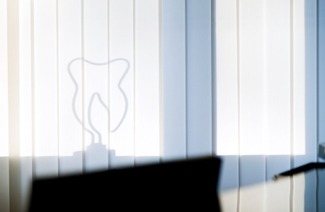 Schatten eines stilisierten Zahns auf vertikalen Lamellen einer Fensterjalousie, im Vordergrund unscharfe Umrisse eines Schreibtisches und einer Lampe.