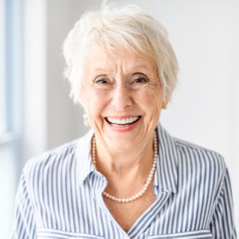 Ältere Frau mit kurzen weißen Haaren lacht herzlich in die Kamera. Sie trägt eine blau-weiß gestreifte Bluse und eine Perlenkette