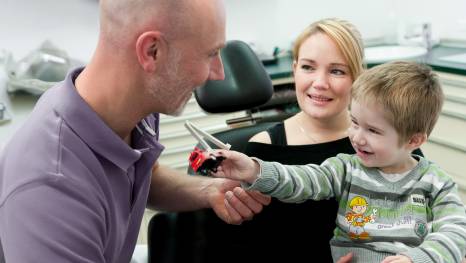 In einer Zahnarztpraxis überreicht ein lachender Junge dem behandelnden Zahnarzt, einem glatzköpfigen Mann in lila Poloshirt, ein Spielzeugauto